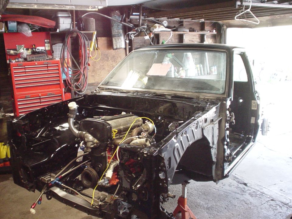 Black pickup under construction in garage, front end removed showing engine with turbo, red toolbox behind
