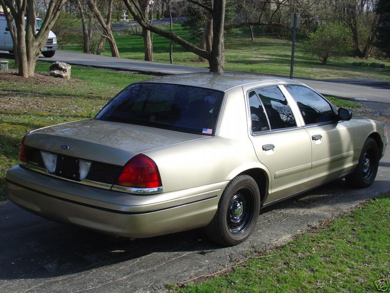Gold Ford Crown Victoria sedan rear three-quarter on a paved driveway with bare trees