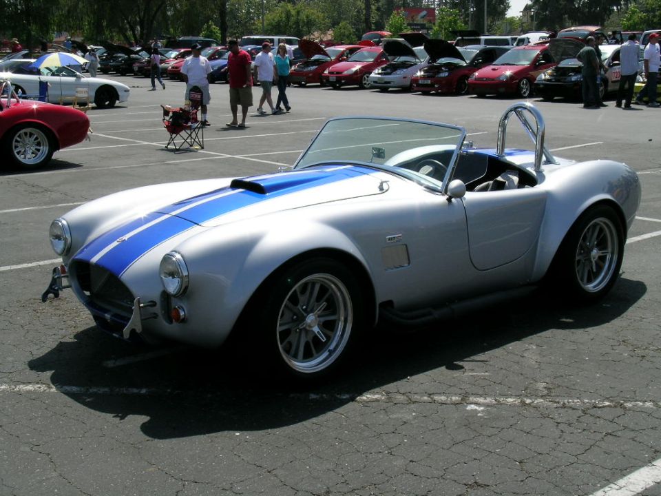 Silver convertible with blue stripes, roll bar, and aftermarket wheels. Other cars and people in background at an outdoor car show