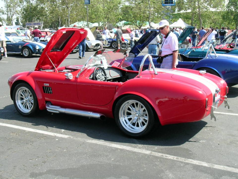 Red convertible sports car with hood open, parked at an outdoor car show. Other cars and people are visible in the background