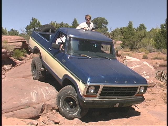 Blue and cream 1978 Ford Bronco crawling down red rocks, passenger standing through roof