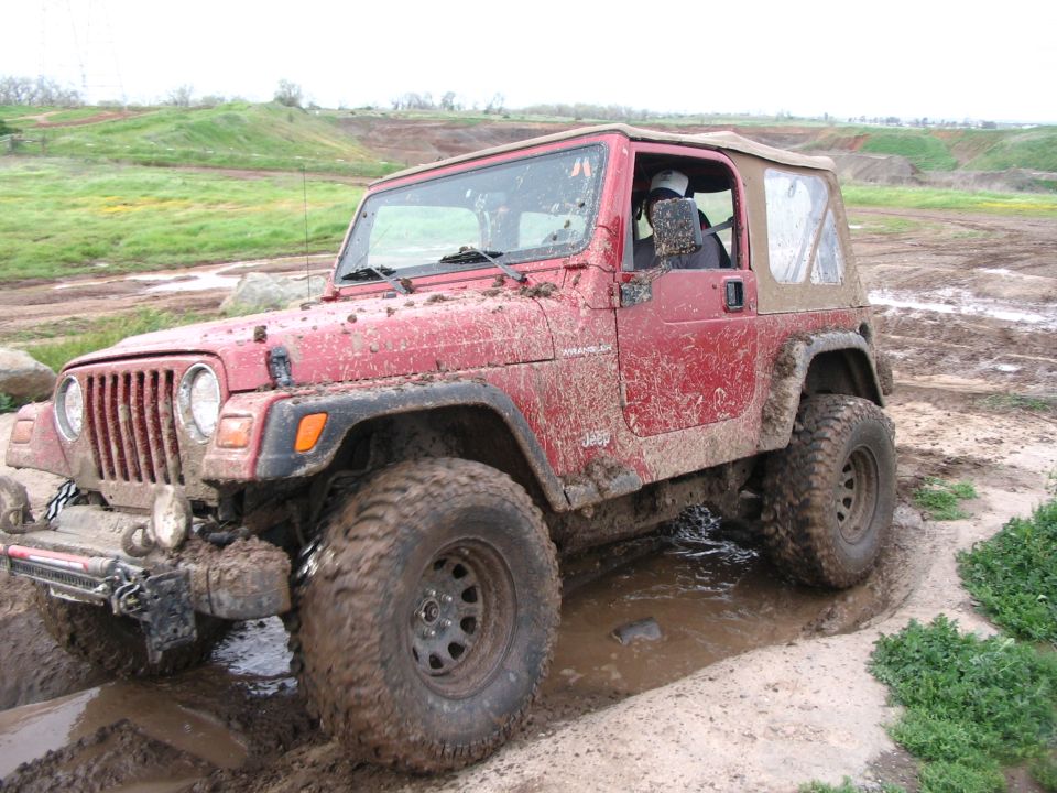 Red Jeep Wrangler with soft top covered in mud crawling through a rutted mud pit