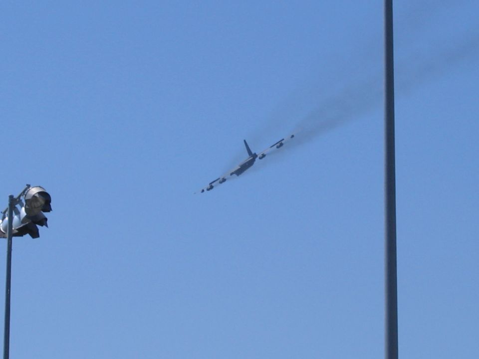 Airplane flying in the sky with smoke trail. A light pole with flags is in the foreground