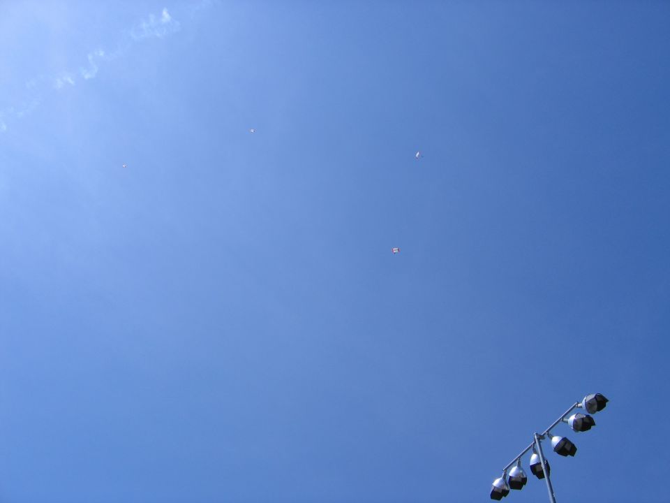 Clear blue sky with tiny distant parachutes and the top of a stadium light tower