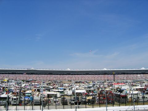 2000 Ford Crown Victoria - A vast parking area filled with vehicles and RVs surrounds a packed racetrack stadium on a clear day.