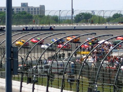 2000 Ford Crown Victoria - Multiple race cars compete on a track, with spectators in the stands and buildings beyond.