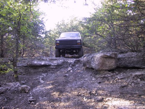 1983 Ford Bronco - A dark SUV navigates a rocky trail through a wooded area.