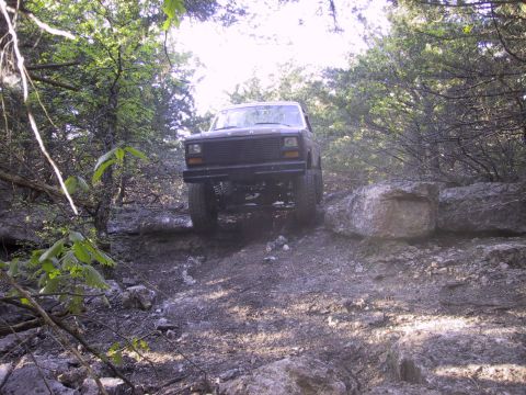 1983 Ford Bronco - This dark SUV is shown from the front as it traverses a challenging rocky path surrounded by trees.
