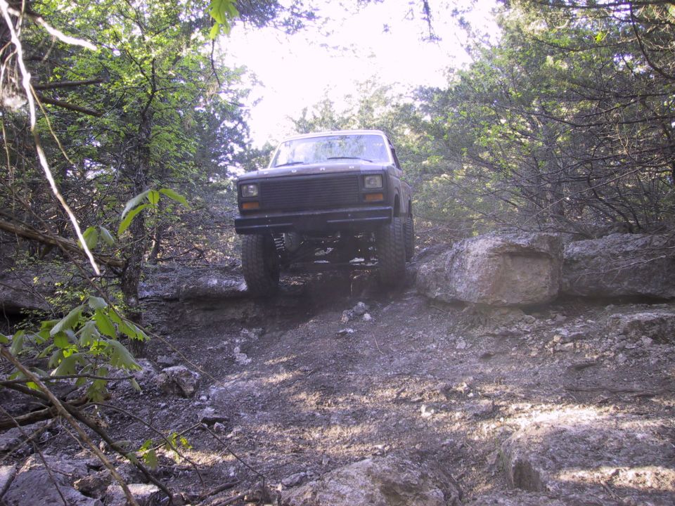 Dark lifted Ford 4x4 cresting a limestone rock ledge on a wooded trail, front axle exposed