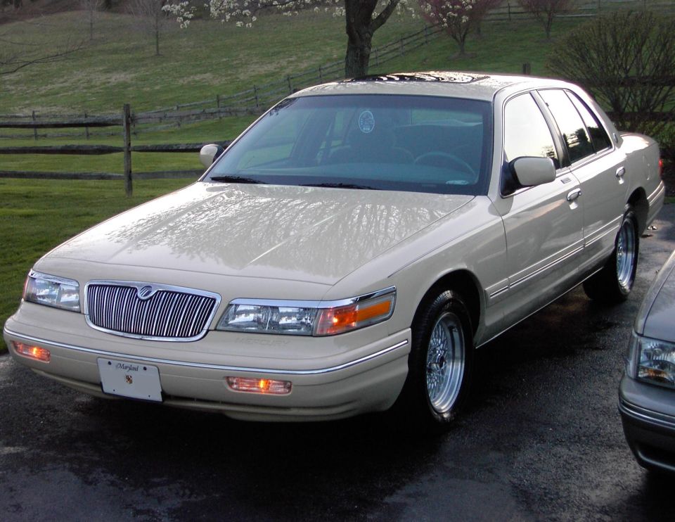 Cream 1996 Mercury Grand Marquis sedan, front three-quarter view, parked on wet pavement near a split-rail fence.