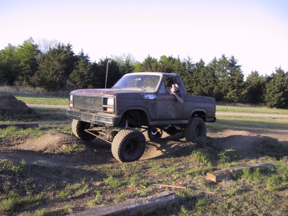 Lifted weathered Ford Bronco climbing a dirt mound in a field, front axle and steering exposed