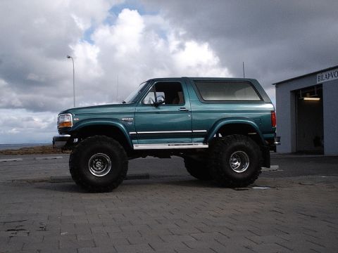 1996 Ford Bronco - A lifted 1996 Ford Bronco with oversized tires is parked outside a building under a cloudy sky.