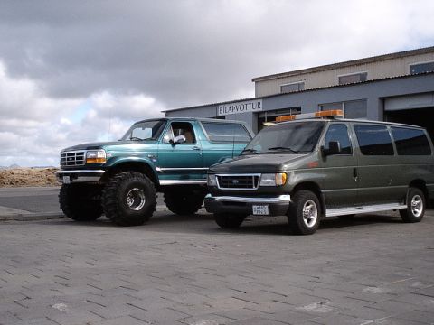 1996 Ford Bronco - Two vehicles, a lifted Ford Bronco and a Ford Econoline van, are parked outside a building with the sign "BILAPVOTTUR".
