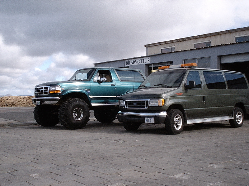 Lifted teal Ford Bronco on big tires next to a green Ford van outside a building