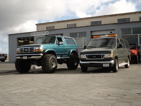 1996 Ford Bronco - A lifted 1996 Ford Bronco with oversized tires is parked outside a building, alongside a dark van and a red Jeep.