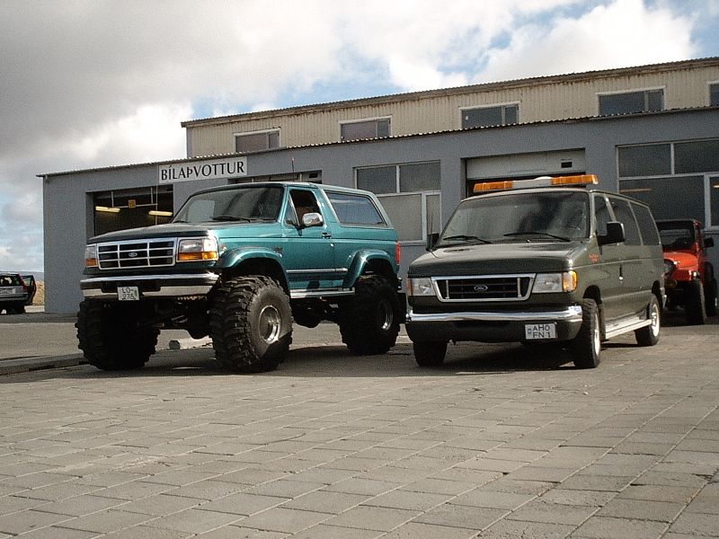 Lifted green Ford Bronco on big tires next to dark green Ford van outside a building