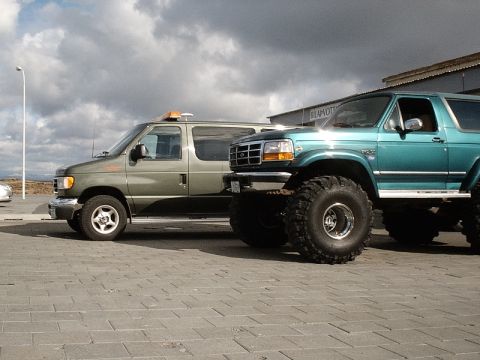 1996 Ford Bronco - A modified 1996 Ford Bronco with large tires is parked next to a green van under a cloudy sky.