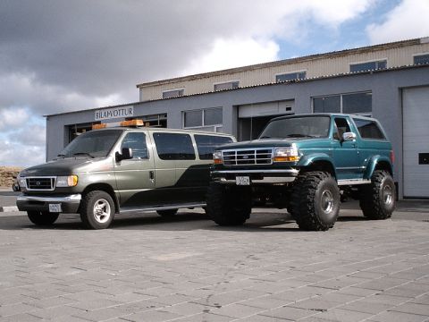 1996 Ford Bronco - A lifted teal Ford Bronco is parked beside a green van in front of a building.