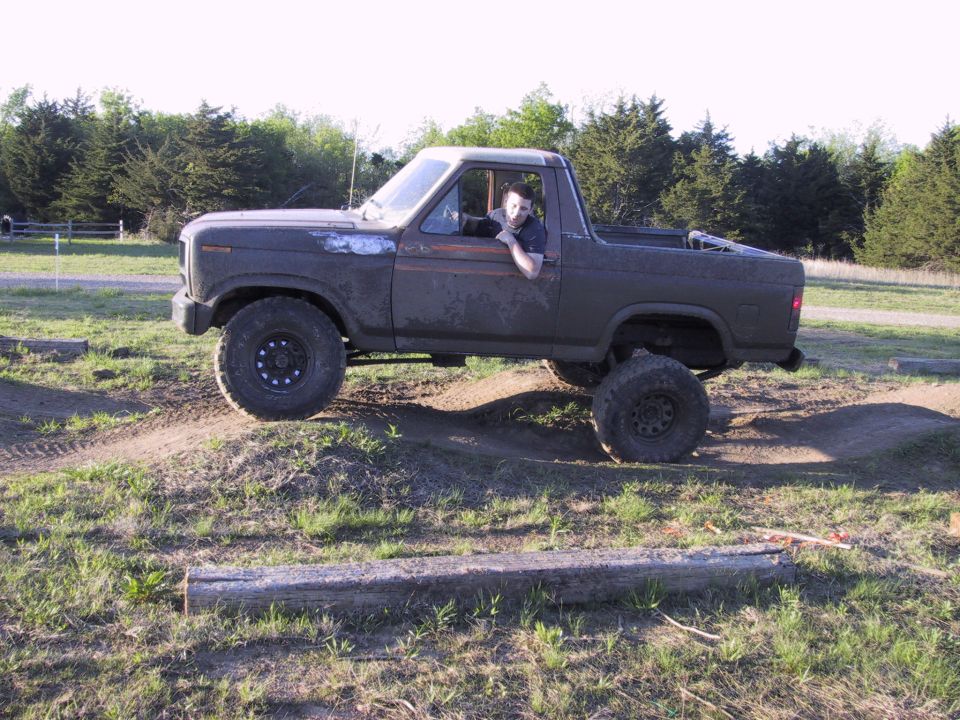 Lifted primer-grey Ford Bronco with rear top removed flexing over dirt mounds, driver in window