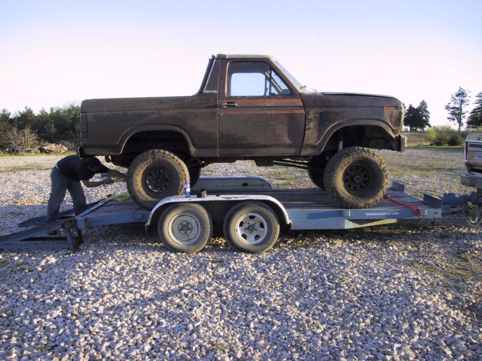 Lifted brown Ford Bronco with top removed loaded on a blue tandem-axle trailer on gravel
