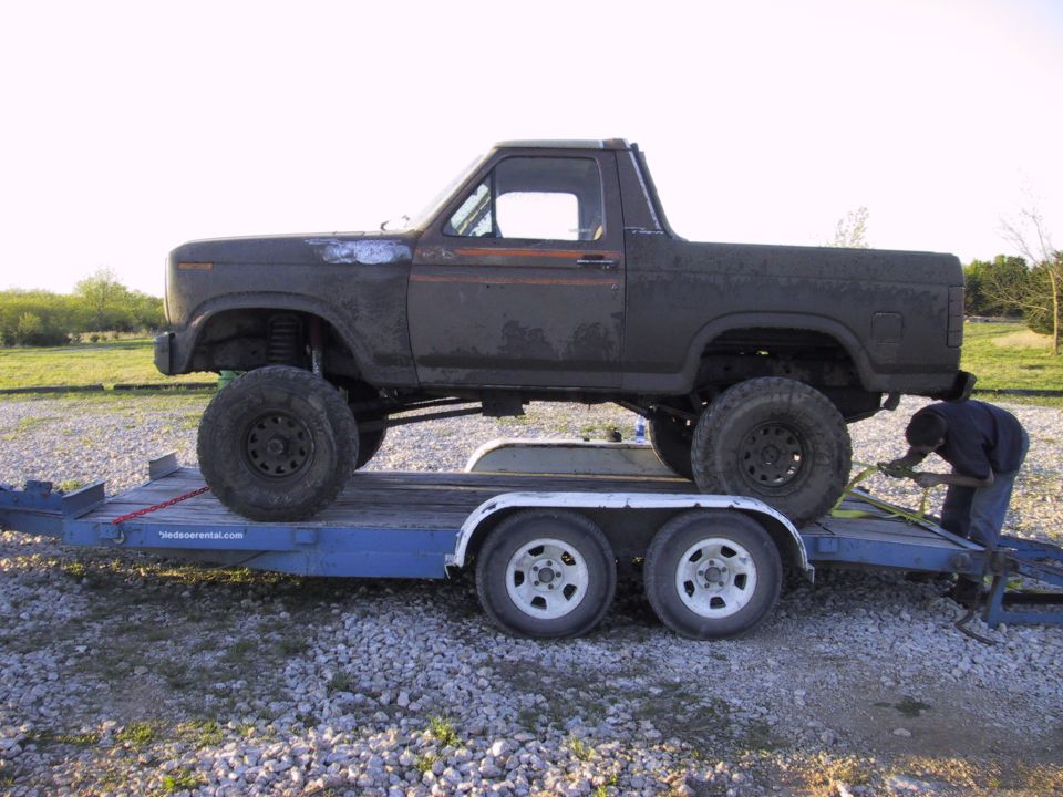 Lifted topless Ford Bronco strapped on a blue tandem-axle rental trailer on gravel