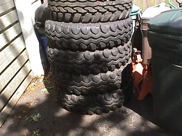 Stack of used off-road mud tires leaning against vinyl siding next to a trash can and jack
