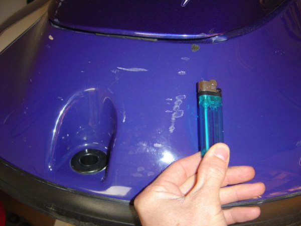 Purple watercraft hull panel with scratches, hand holding a lighter for scale near a plug hole.