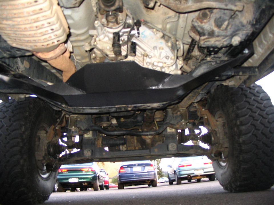Underbody view of a lifted truck showing transmission crossmember and driveshaft over a parking lot