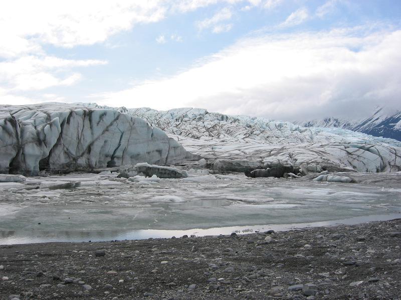 Wide glacier flowing down toward a gravel shore with mountains behind