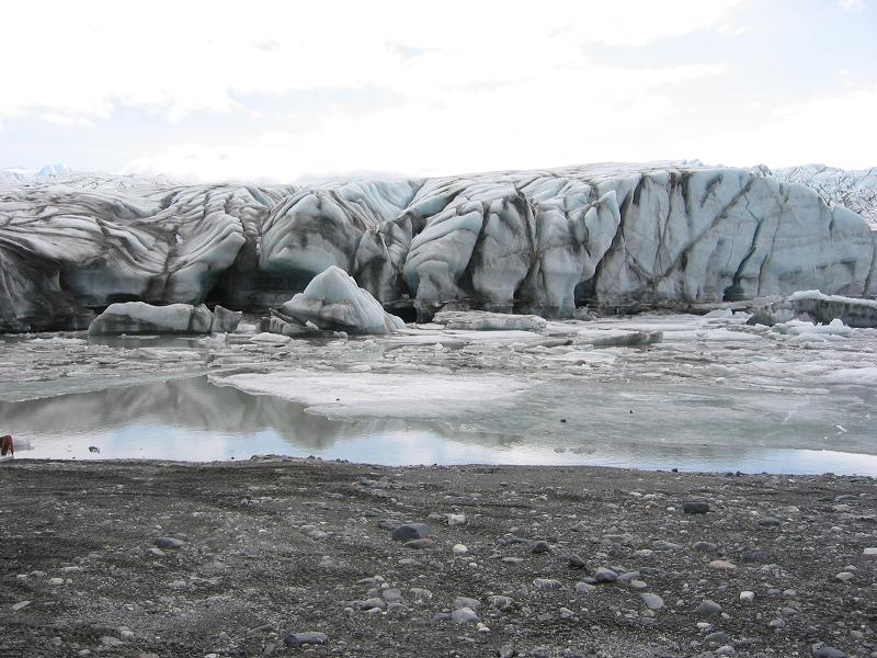 Glacier terminus and ice-choked pond seen across a dark gravel beach