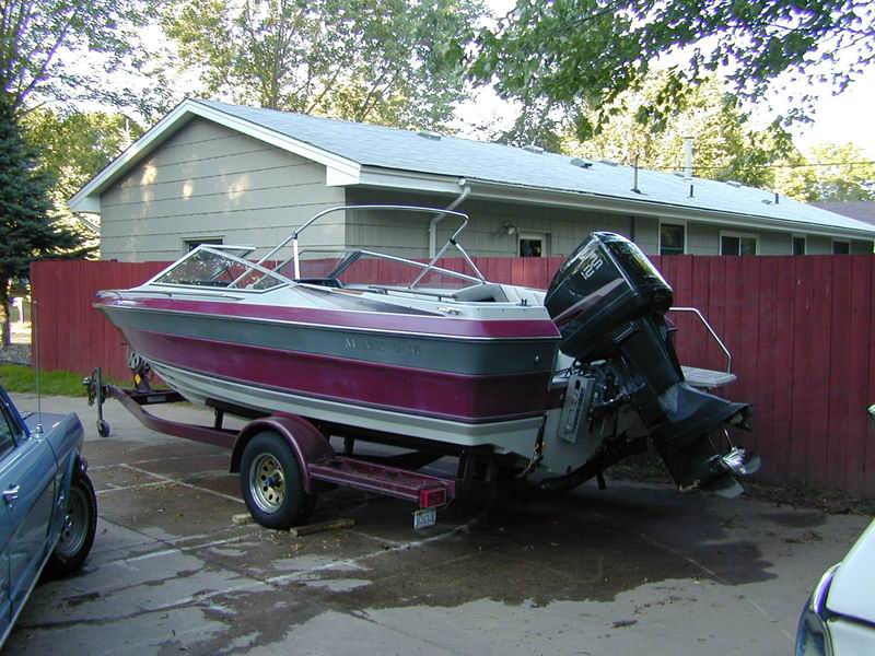 Purple and white runabout boat on trailer, rear-left view with outboard motor, parked by a fence