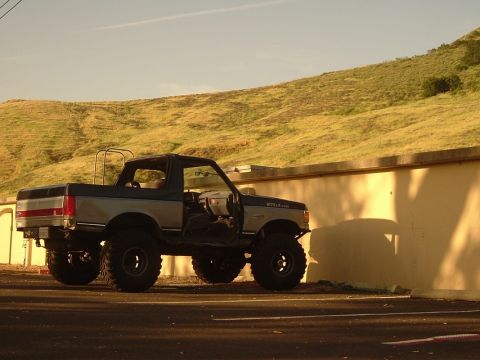 1989 Ford Bronco - This lifted Ford Bronco, likely from the late 1980s, is parked beside a textured wall under a bright sky.