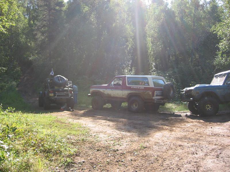 Lifted red and white Ford Bronco on mud tires parked in a sunlit forest trail clearing