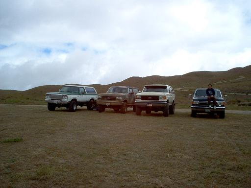 Four lifted 4x4 trucks parked in a row in a grassy field under overcast sky