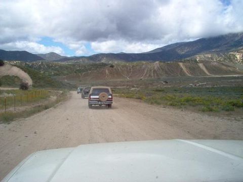 1988 Ford Bronco - A convoy of three SUVs, including a 1988 Ford Bronco, travels down a dusty mountain road under a cloudy sky.