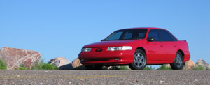 1995 Ford Taurus - A red 1995 Ford Taurus is parked on a gravel road under a clear blue sky.