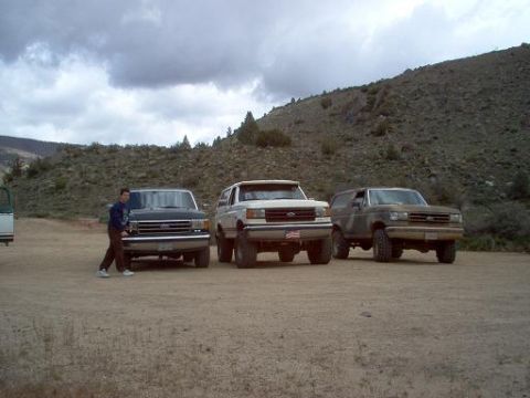 1988 Ford Bronco - Three SUVs are parked in a dusty lot with a man standing by the leftmost vehicle.