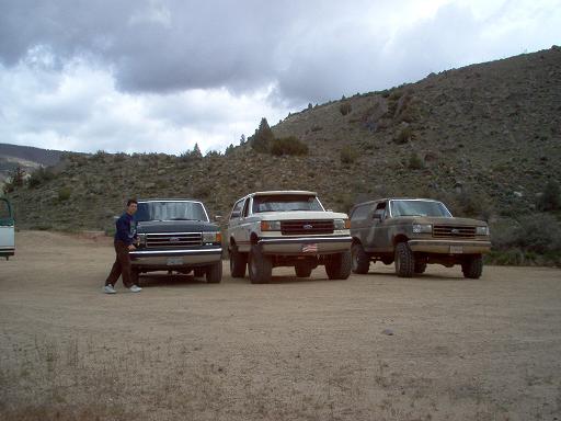 Three SUVs parked on a dirt lot, two are Ford Broncos from the late 1980s, one is a Ford F-Series truck from the late 1980s. A man stands by the truck