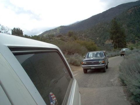 1988 Ford Bronco - A dark blue SUV drives down a rural road, with another vehicle following in the distance.