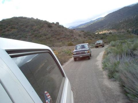 1988 Ford Bronco - Two trucks travel on a paved road through a mountainous, arid landscape under a cloudy sky.