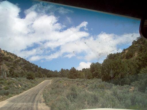 View from a vehicle looking down a dirt road through hills and trees under a cloudy sky