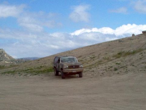 1988 Ford Bronco - A brown 1988 Ford Bronco is parked on a dusty, barren hillside with a bright blue sky overhead.