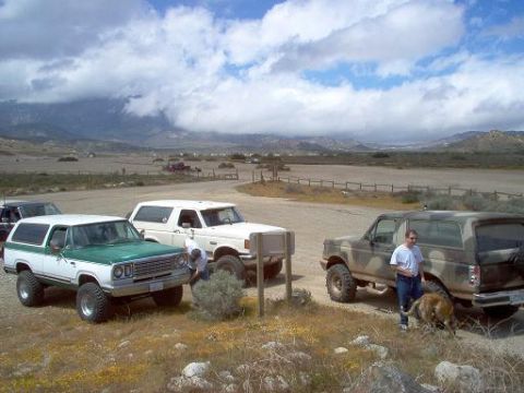 1988 Ford Bronco - Three SUVs are parked in a dry, open area with mountains in the background. A man and dog are near one of the vehicles.