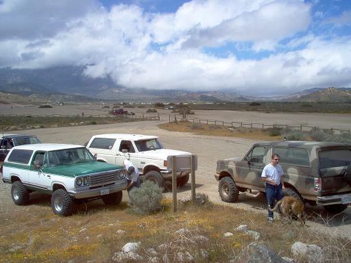 Three SUVs parked in a dry, mountainous area under a cloudy sky. A man and dog stand by a camouflage SUV