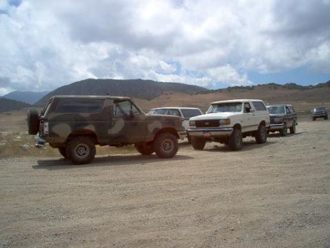 1988 Ford Bronco - Several Ford Bronco SUVs are parked on a dirt road with hills and clouds in the background.