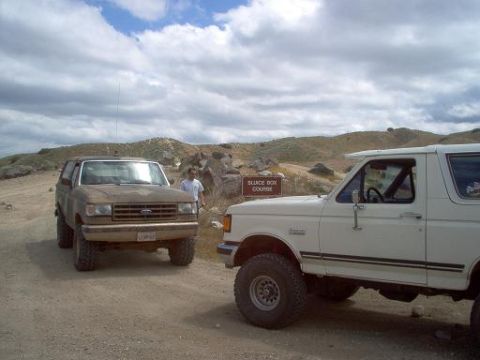 1988 Ford Bronco - Two Ford Broncos are parked on a dirt road with hills in the background. A sign reads 'SLUICE BOX COURSE'.