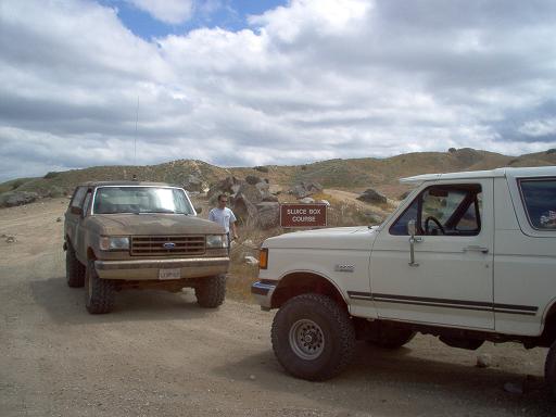 Two lifted Ford Broncos parked at a dirt trailhead beside a Sluice Box Course sign