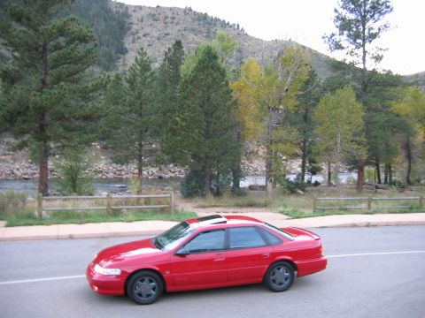 1995 Ford Taurus - A red sedan is parked next to a river with trees and a mountain in the background.