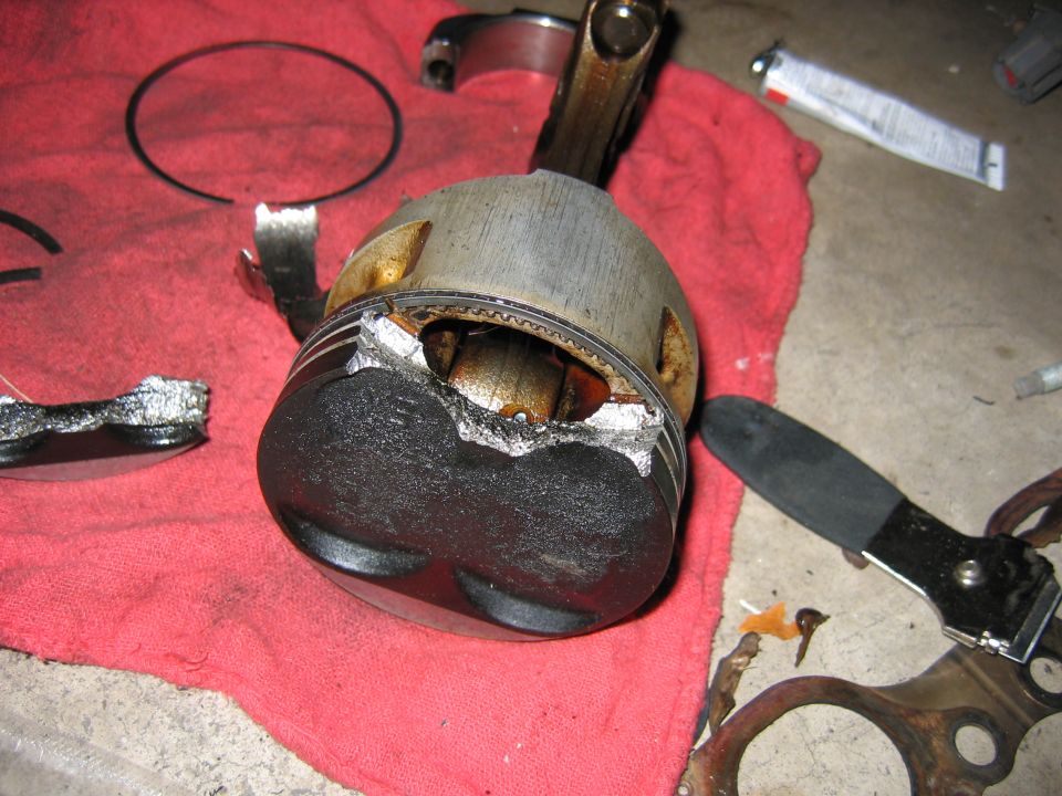Damaged piston with connecting rod attached, lying on a red cloth. Broken piston rings are nearby