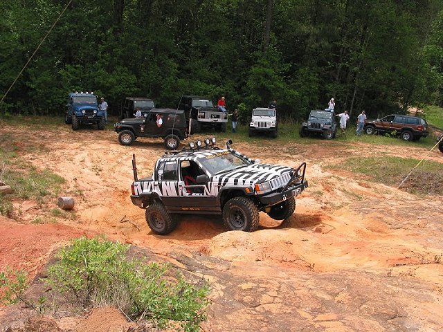 White zebra-striped lifted Jeep SUV climbing dirt trail with other Jeeps parked behind